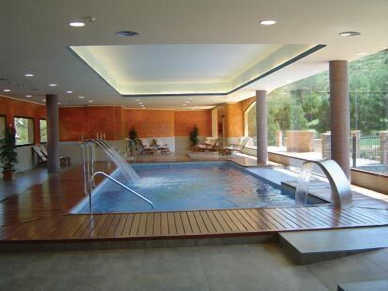 Una piscina interior con cubierta de madera, sillones de descanso y techo de cristal.