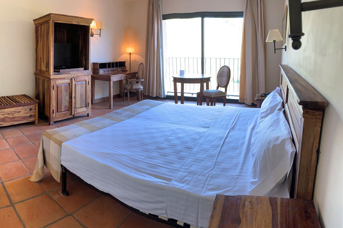 A hotel room with a large bed, a wooden cabinet, a desk with a chair, and a balcony with a view.