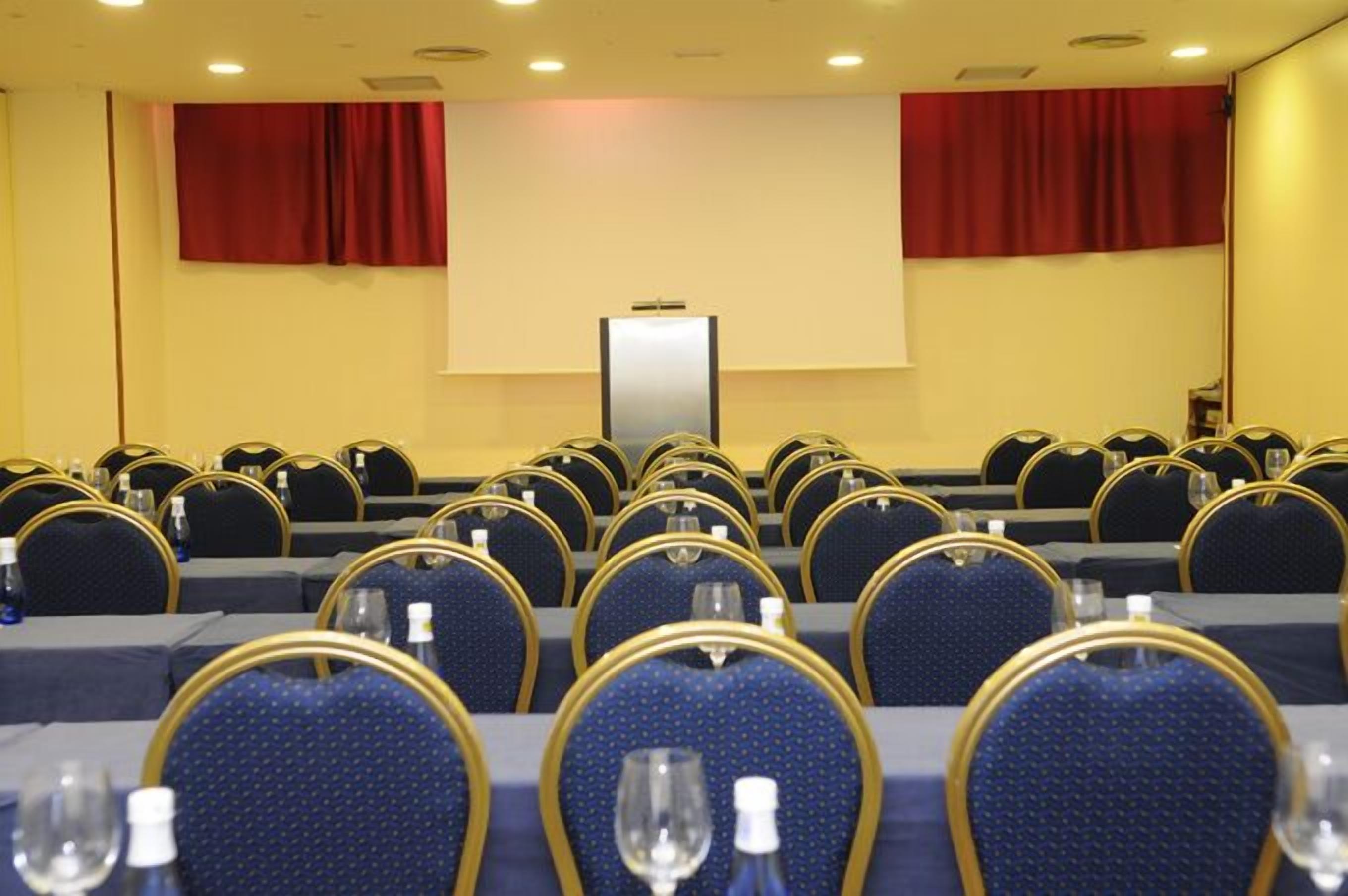 Een congreszaal met rijen blauwe stoelen, een spreekgestoelte en een groot scherm.