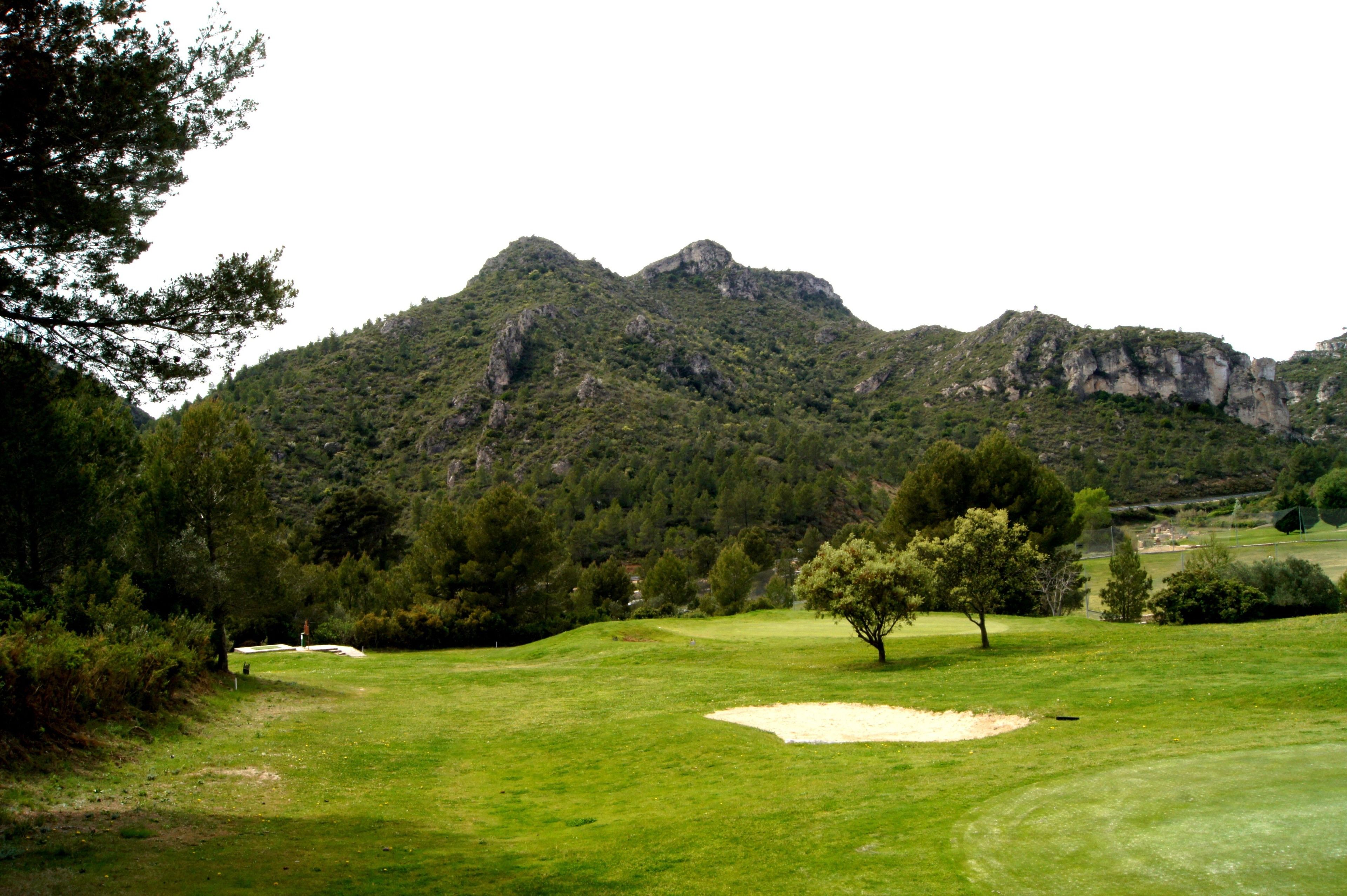 Un campo de golf con un búnker de arena, árboles y montañas al fondo.