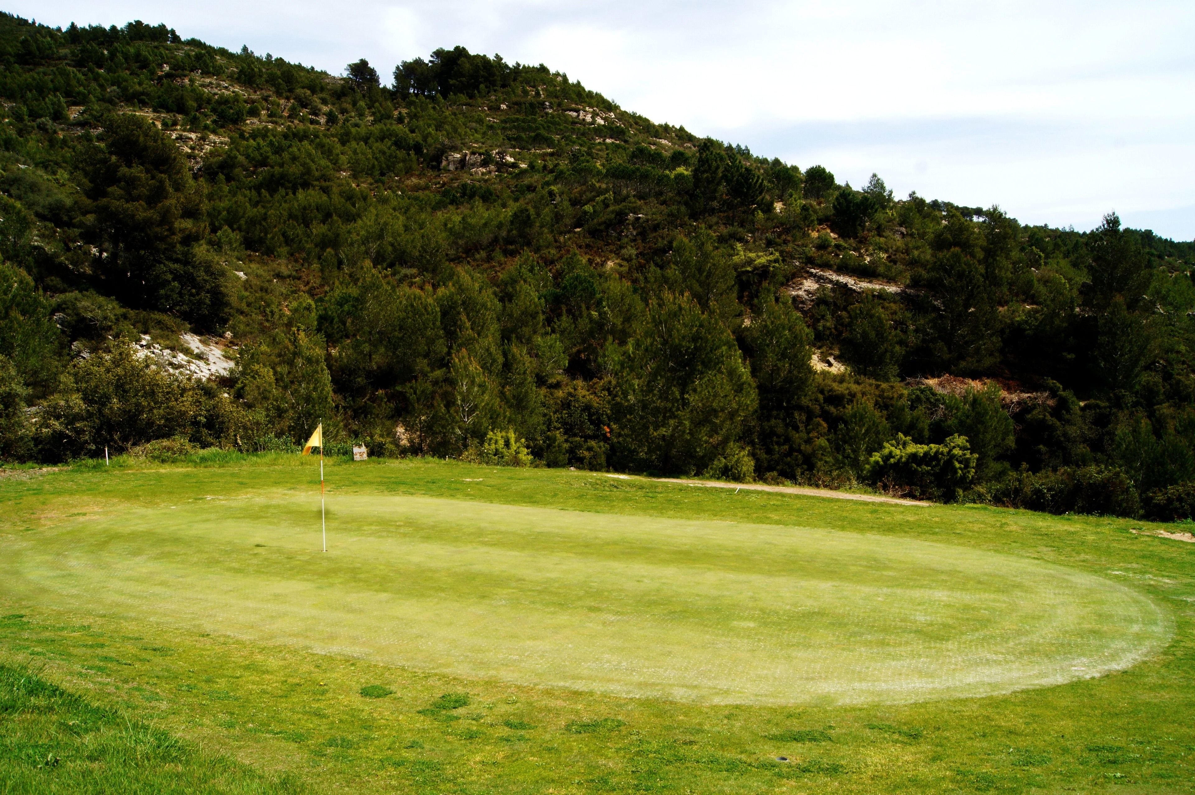 Um campo de golfe com bandeira no green, rodeado por floresta densa e colinas.