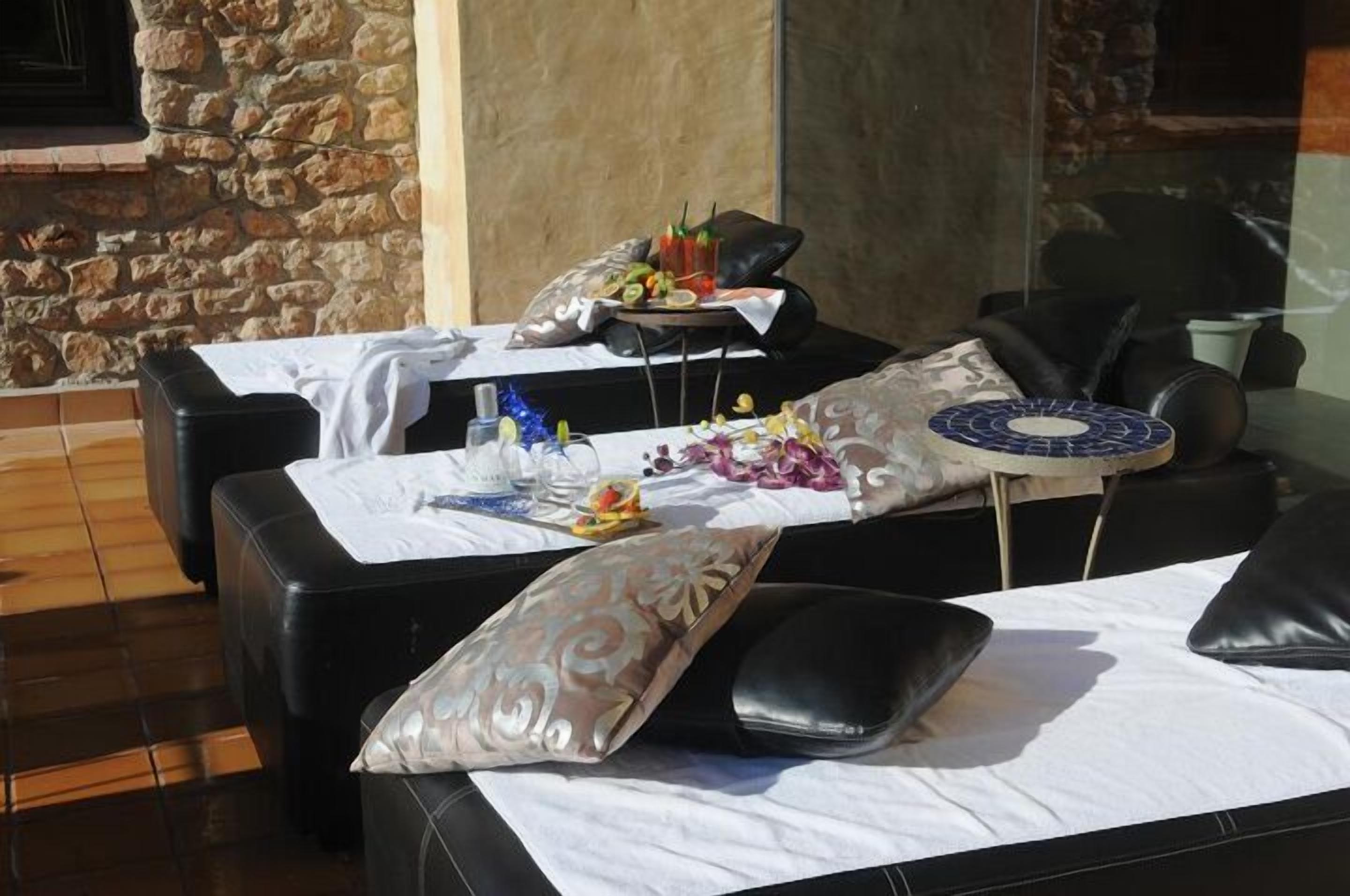 A sunlit outdoor seating area with stone walls, tables with white tablecloths, and black leather chairs.