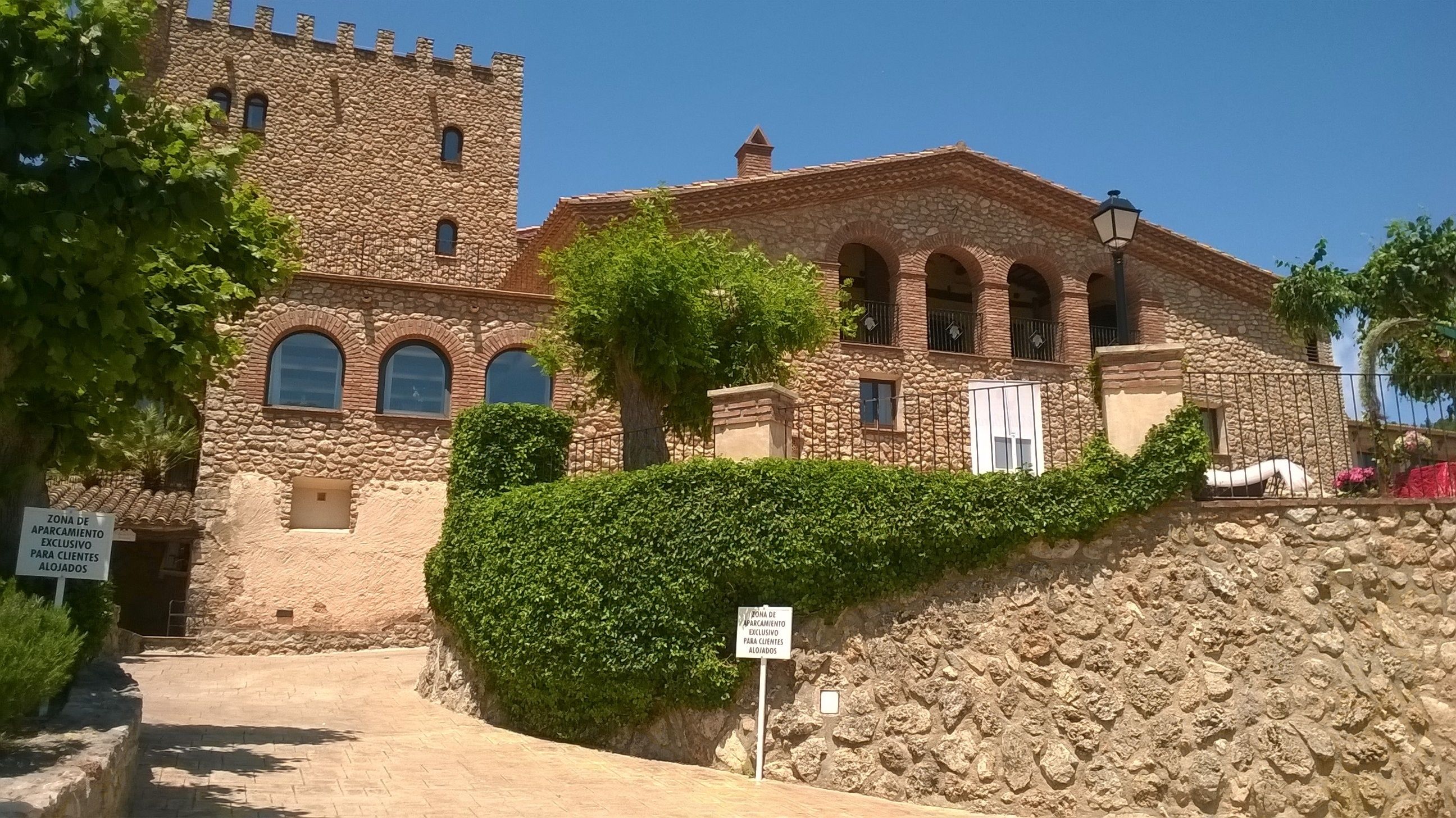 Ein Gebäude aus Stein mit Bogenfenstern und einem Turm, umgeben von Grünflächen und einem gepflasterten Eingang.