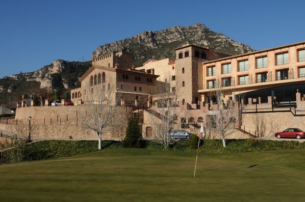 Un bâtiment qui ressemble à un château, avec une façade en pierre, une grande pelouse devant et une montagne en arrière-plan.