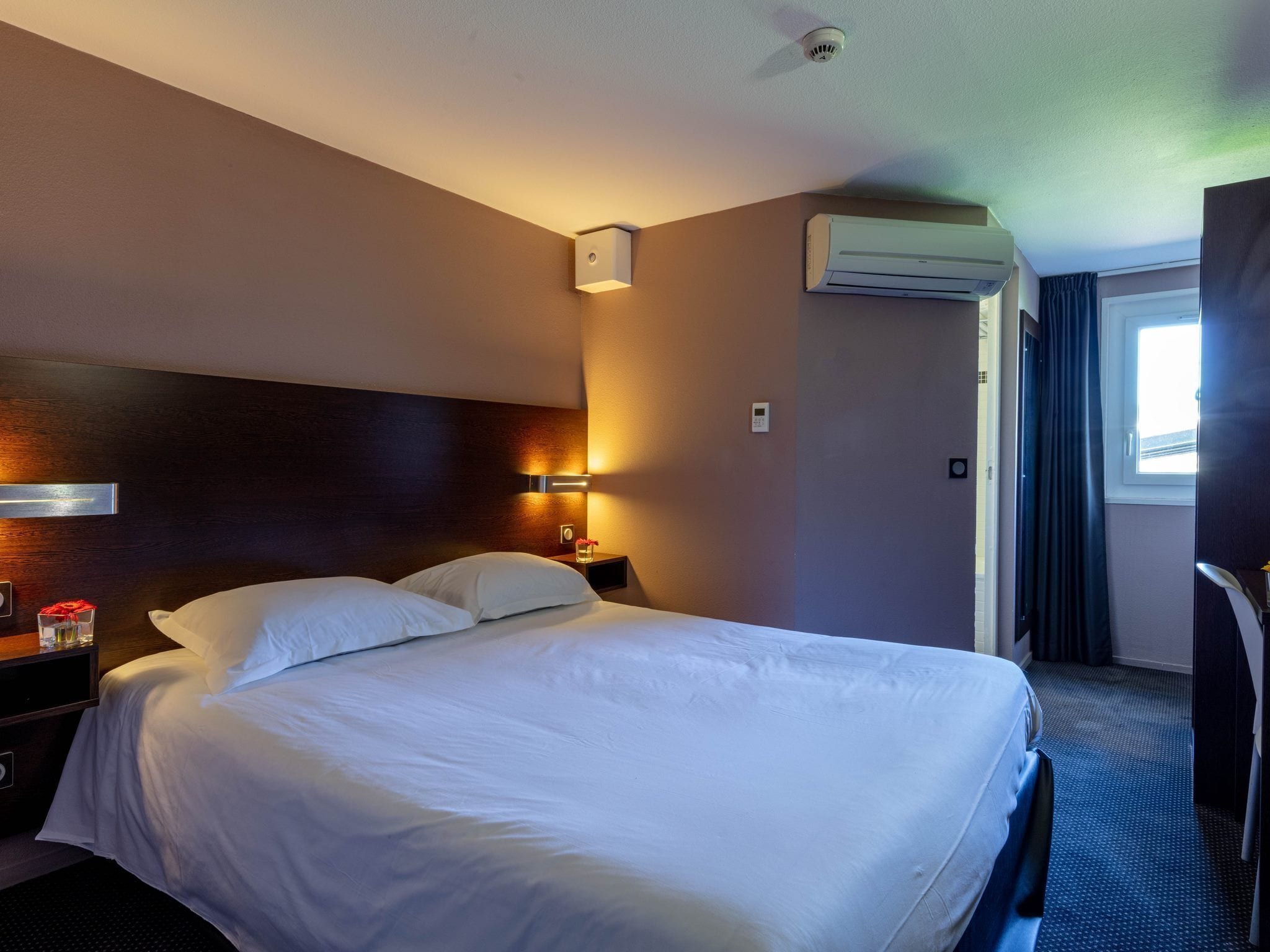 A hotel room with a large bed, a bedside table with a lamp, an air conditioning unit, and a window with curtains.