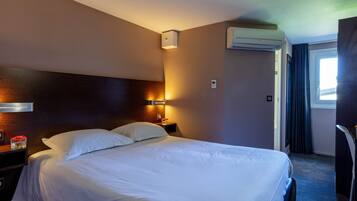 A hotel room with a large bed, a bedside table with a lamp, an air conditioning unit, and a window with curtains.
