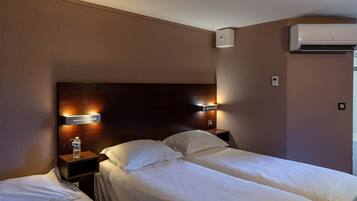 Een hotelkamer met twee bedden, een houten hoofdbord, wandlampen, een waterfles op het nachtkastje en een airconditioner.