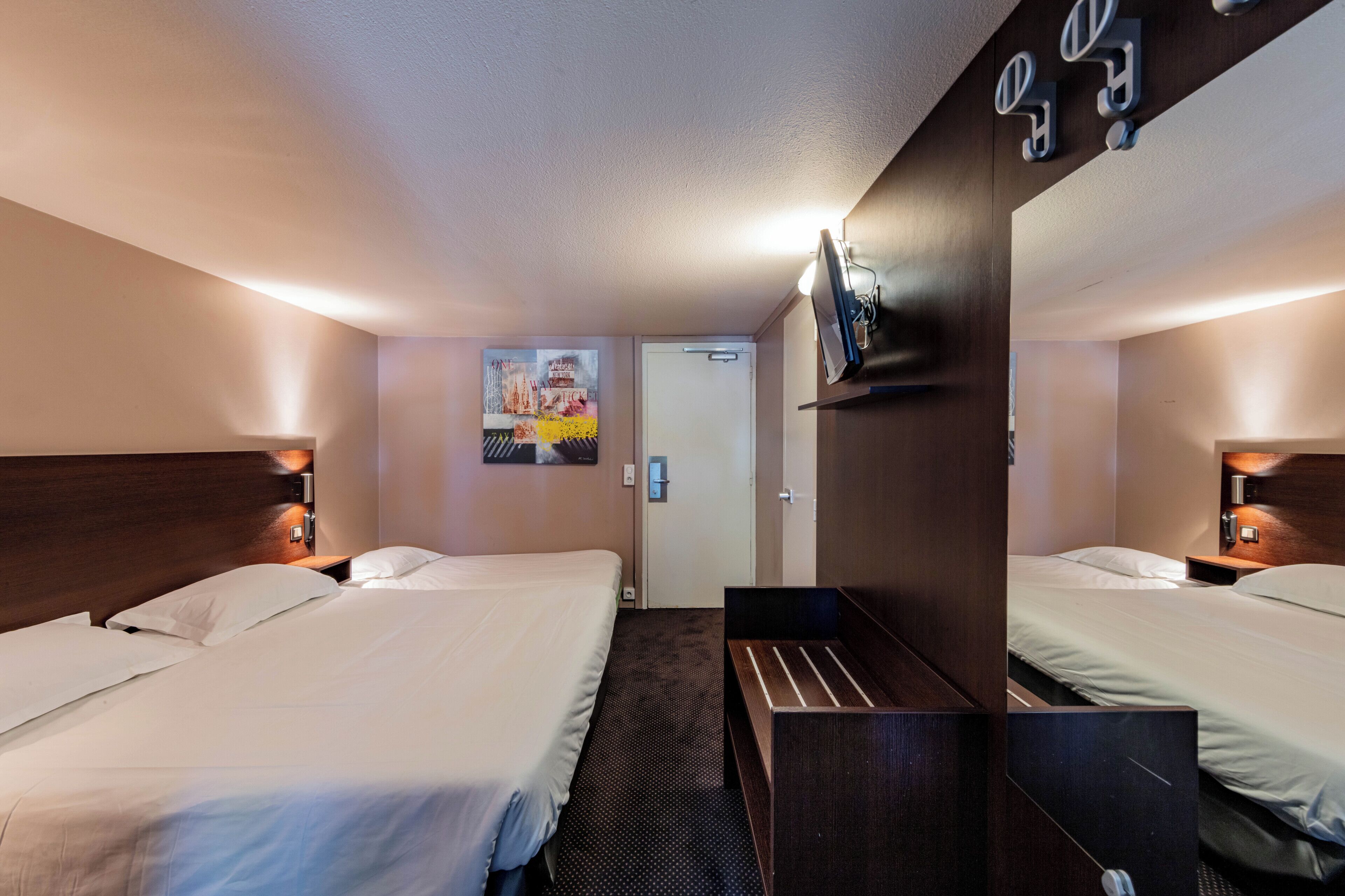 Habitación de hotel con dos camas, un escritorio pequeño, un espejo y un televisor montado en la pared.