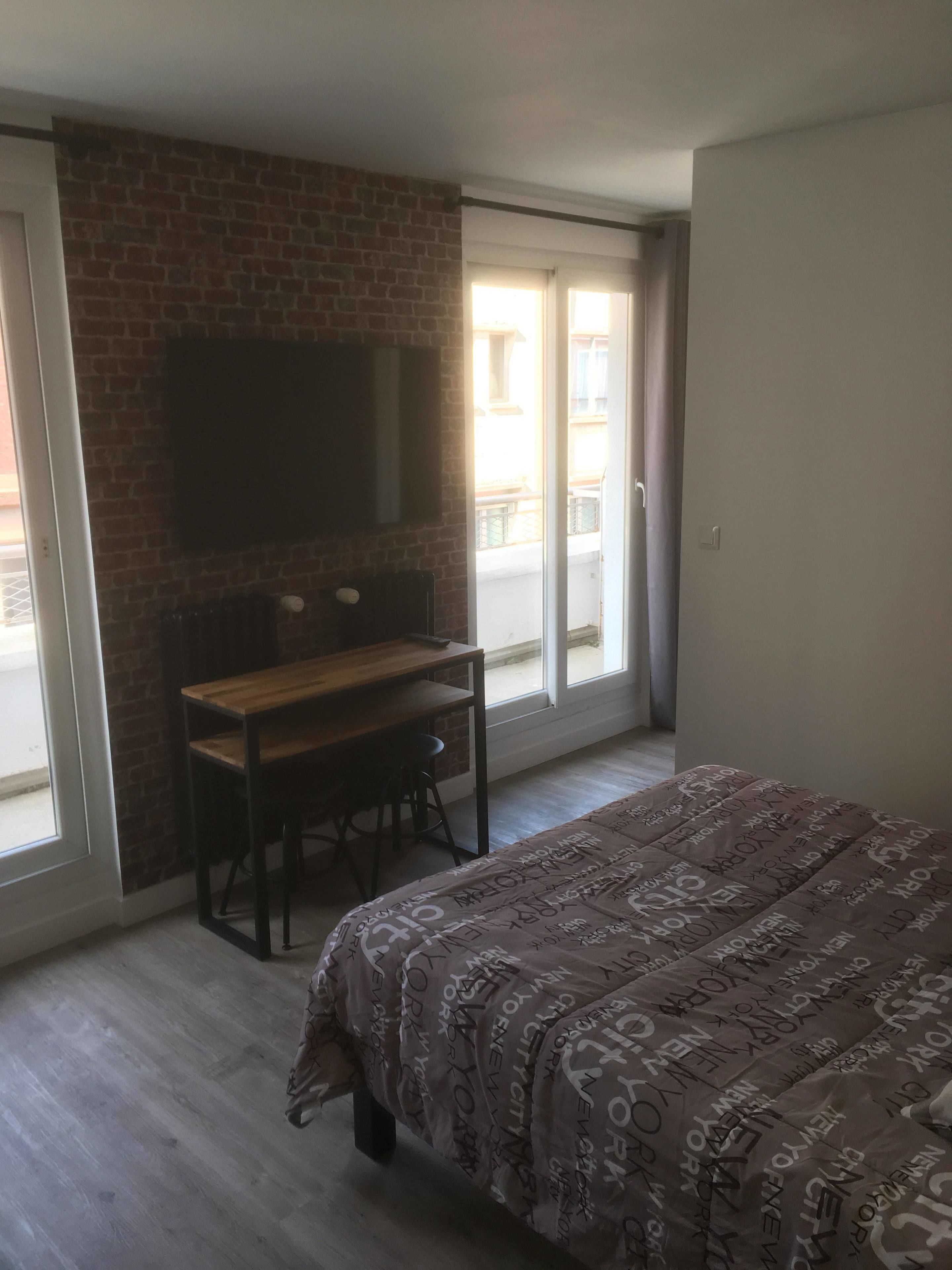 A bedroom with a bed, a desk, and a TV mounted on a brick wall.