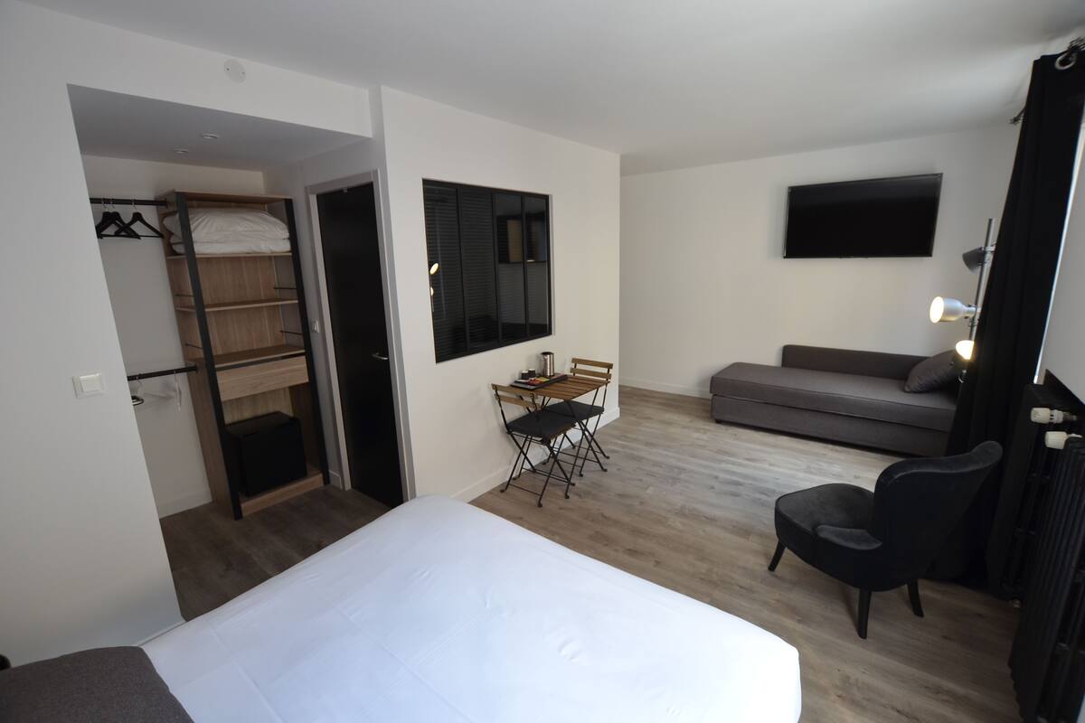 A hotel room with a bed, a desk with a chair, a sofa, a TV mounted on the wall, and a wardrobe with a mirror.