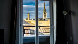 Vista desde un balcón con ventana, que muestra un edificio con una aguja alta y otra estructura con chimenea.