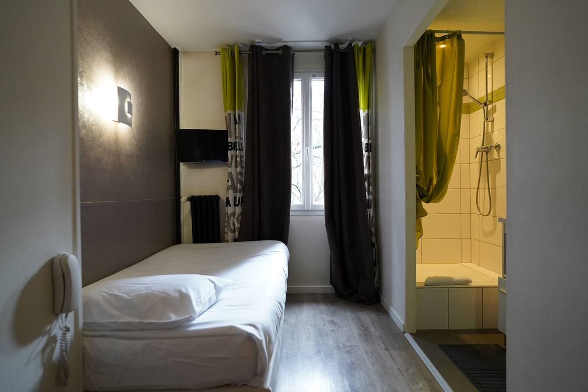 A small, compact room with a single bed, a shower area, and a mounted TV.