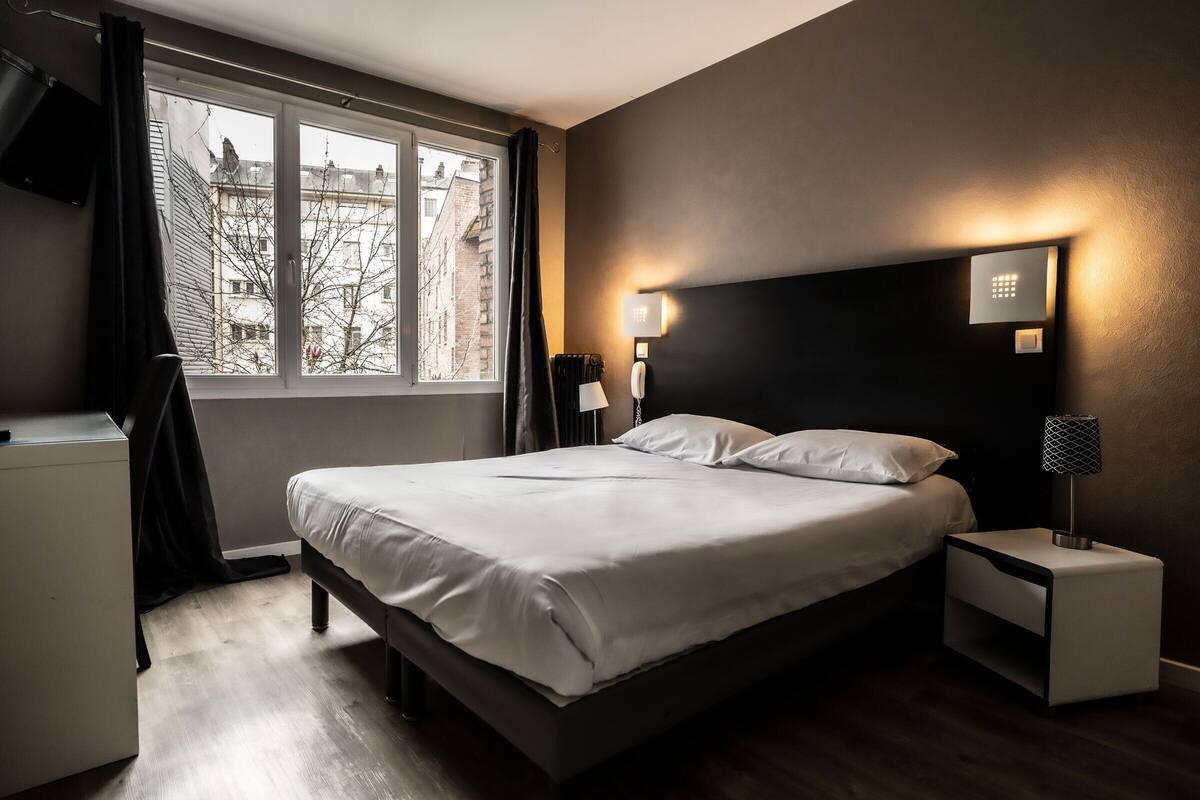 A modern bedroom with a large bed, bedside tables, a window with curtains, and a view of buildings outside.
