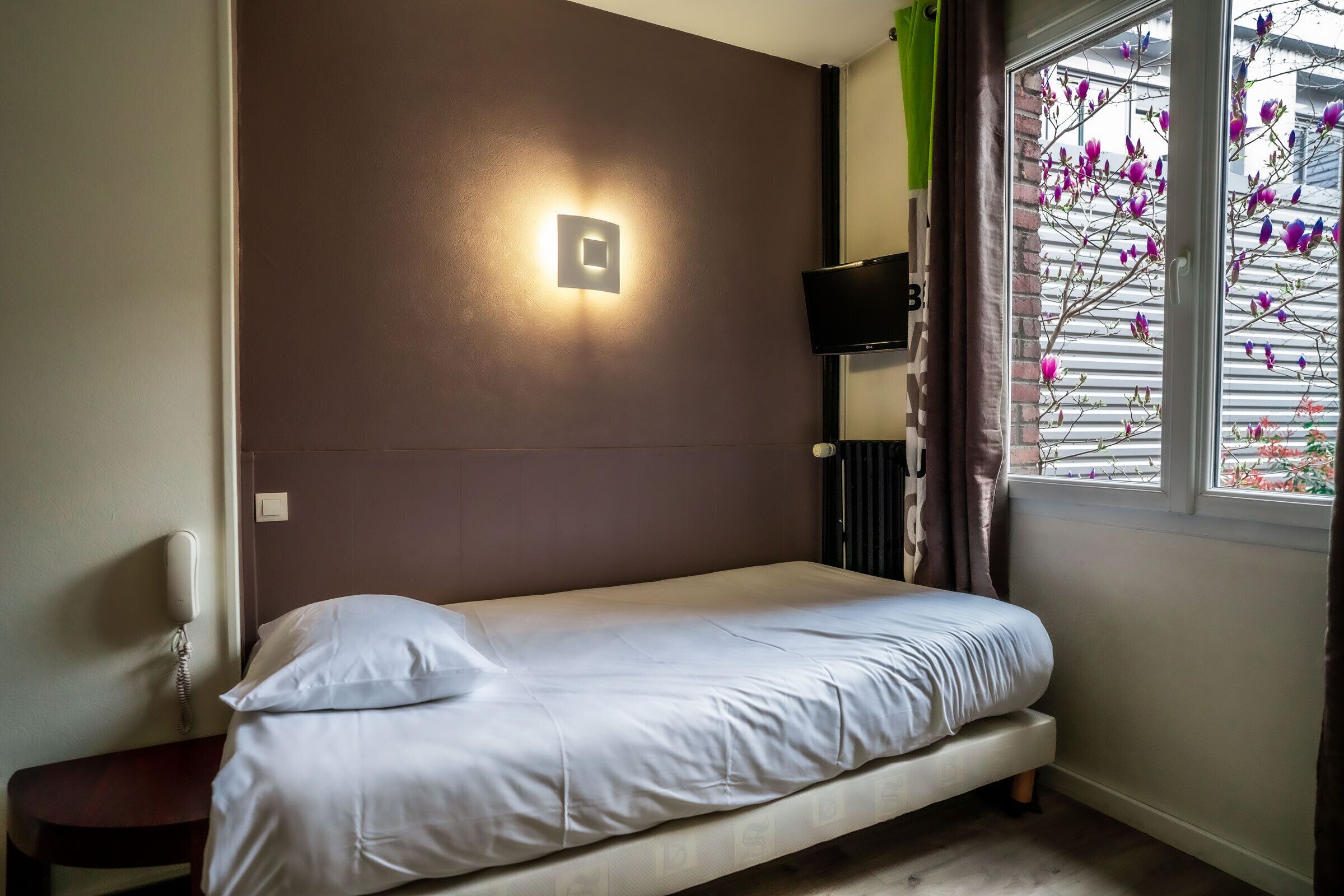 A single bed with a headboard, a bedside table, a wall-mounted TV, and a window with curtains.