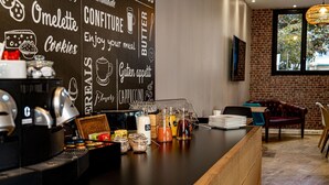 Una cafetería con un menú en una pizarra que enumera opciones como tortilla, mermelada, mantequilla, galletas, capuchino y sabores. Hay máquinas de café, un dispensador de leche y una variedad de aderezos en la barra.
