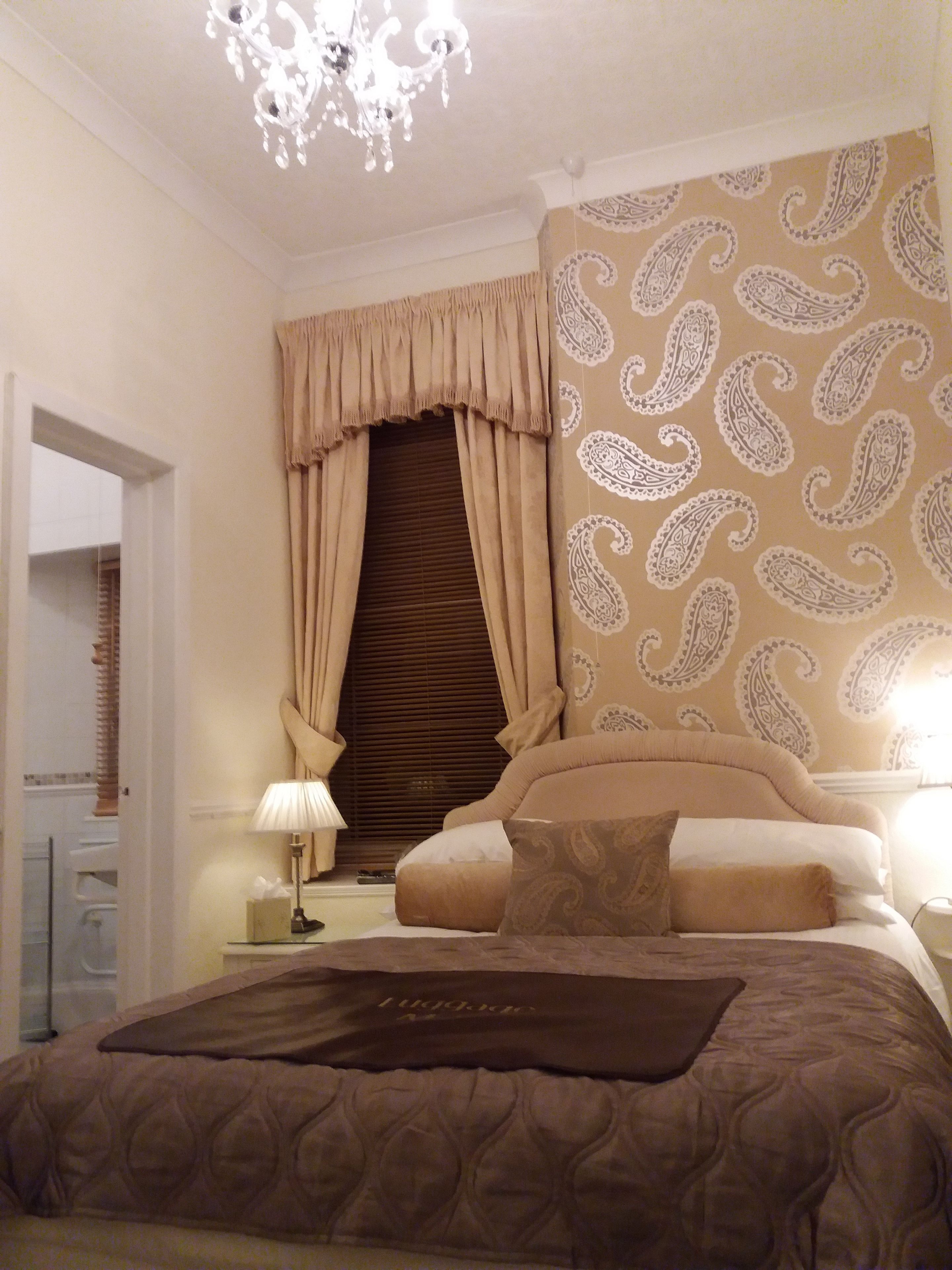 A bedroom with a large bed, a chandelier, a window with curtains, and a patterned wall.