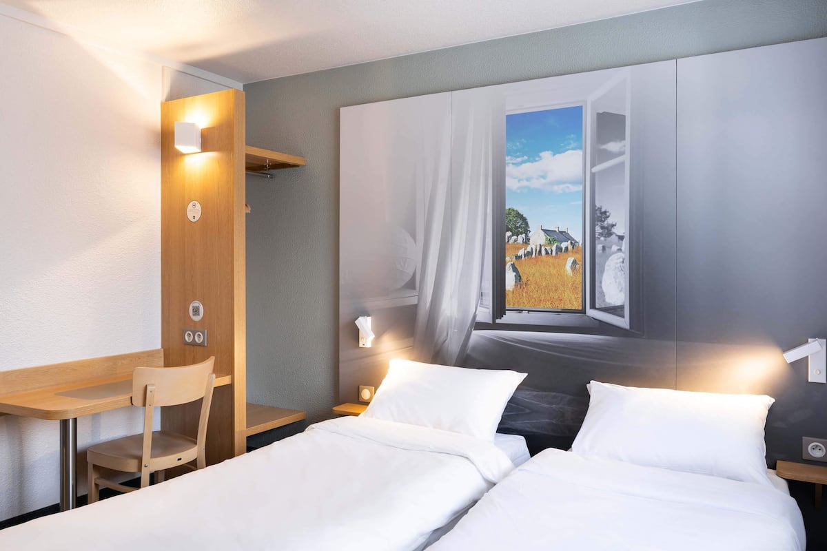 Habitación de hotel con cama, escritorio y silla. Un cuadro en la pared muestra una ventana con vistas a un campo y árboles.