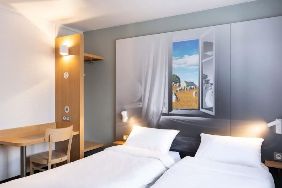 Une chambre d’hôtel avec deux lits, un bureau et une chaise. Un tableau représentant un paysage est accroché au mur.
