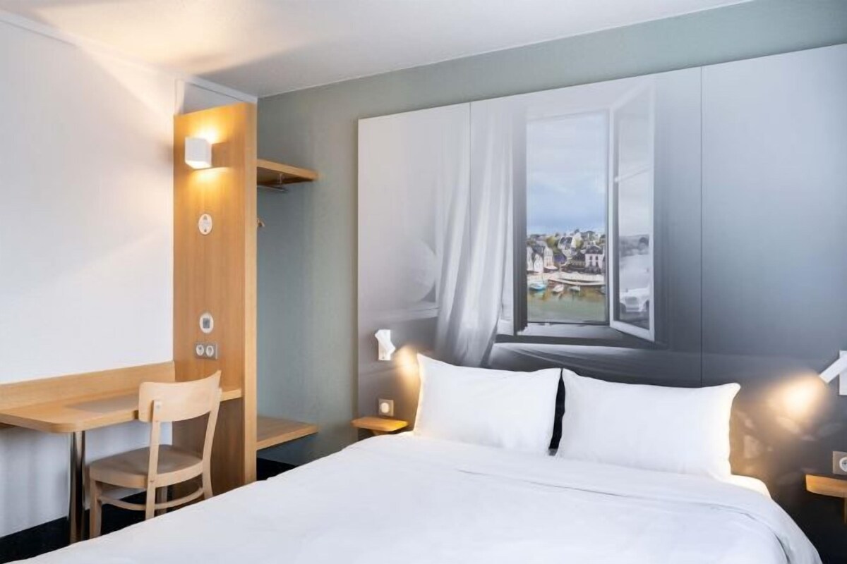 A hotel room with a bed, a desk, and a chair. There is a painting on the wall depicting a window with a view of a cityscape.