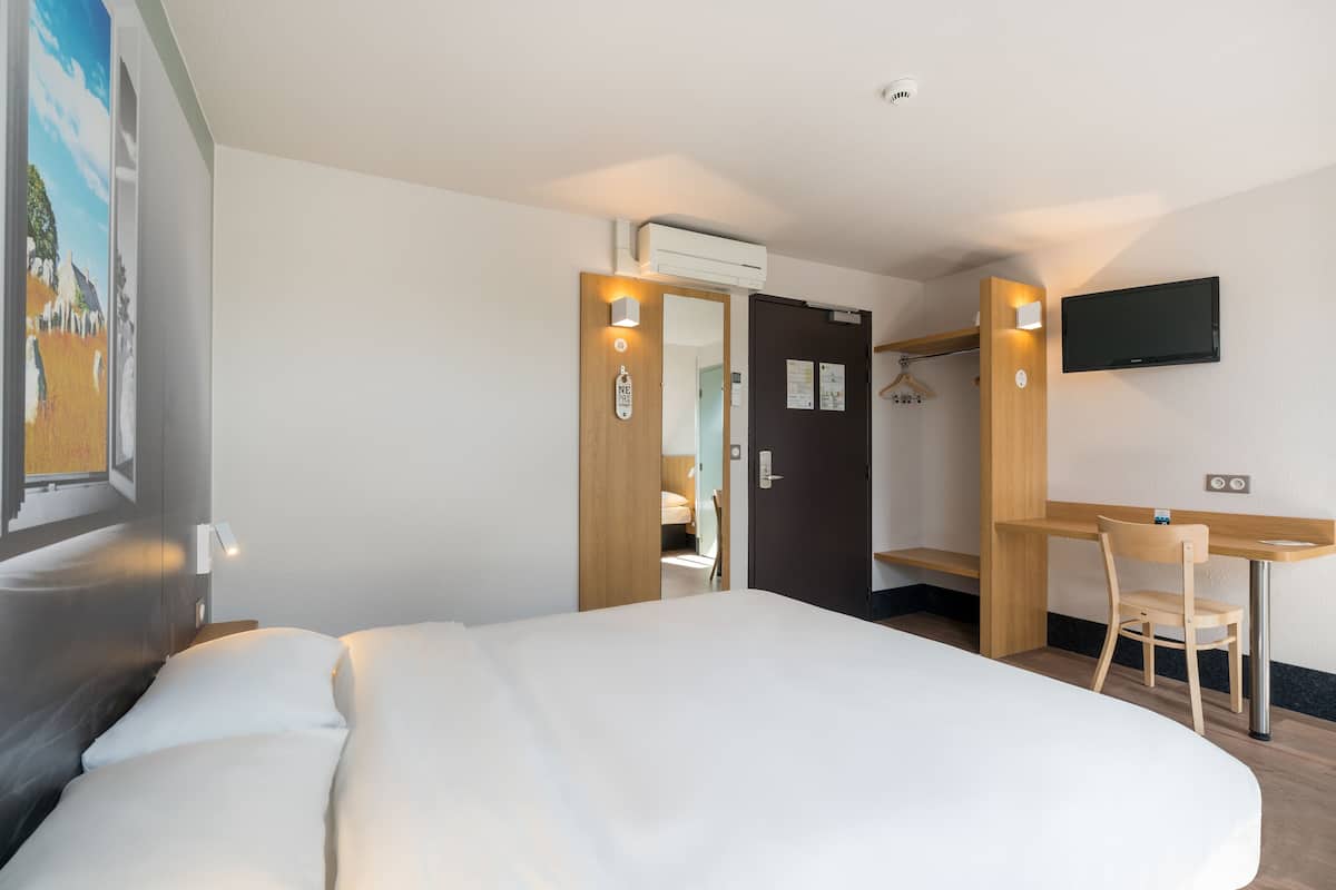 Una habitación de hotel con cama, escritorio, silla, televisor montado en la pared y puerta que da a otra habitación.