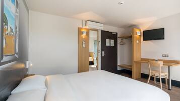 Una habitación de hotel con cama, escritorio, silla, televisor montado en la pared y puerta que da a otra habitación.