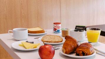 Una mesa de desayuno con tostadas, un croissant, una manzana, un vaso de zumo de naranja, una taza de café, un tarro de mermelada y una bolsita de té.