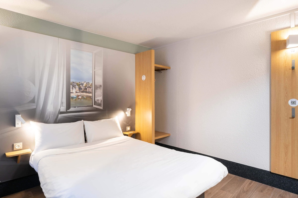 Habitación de hotel con cama, mesitas de noche, estante en la pared y una puerta con código QR.