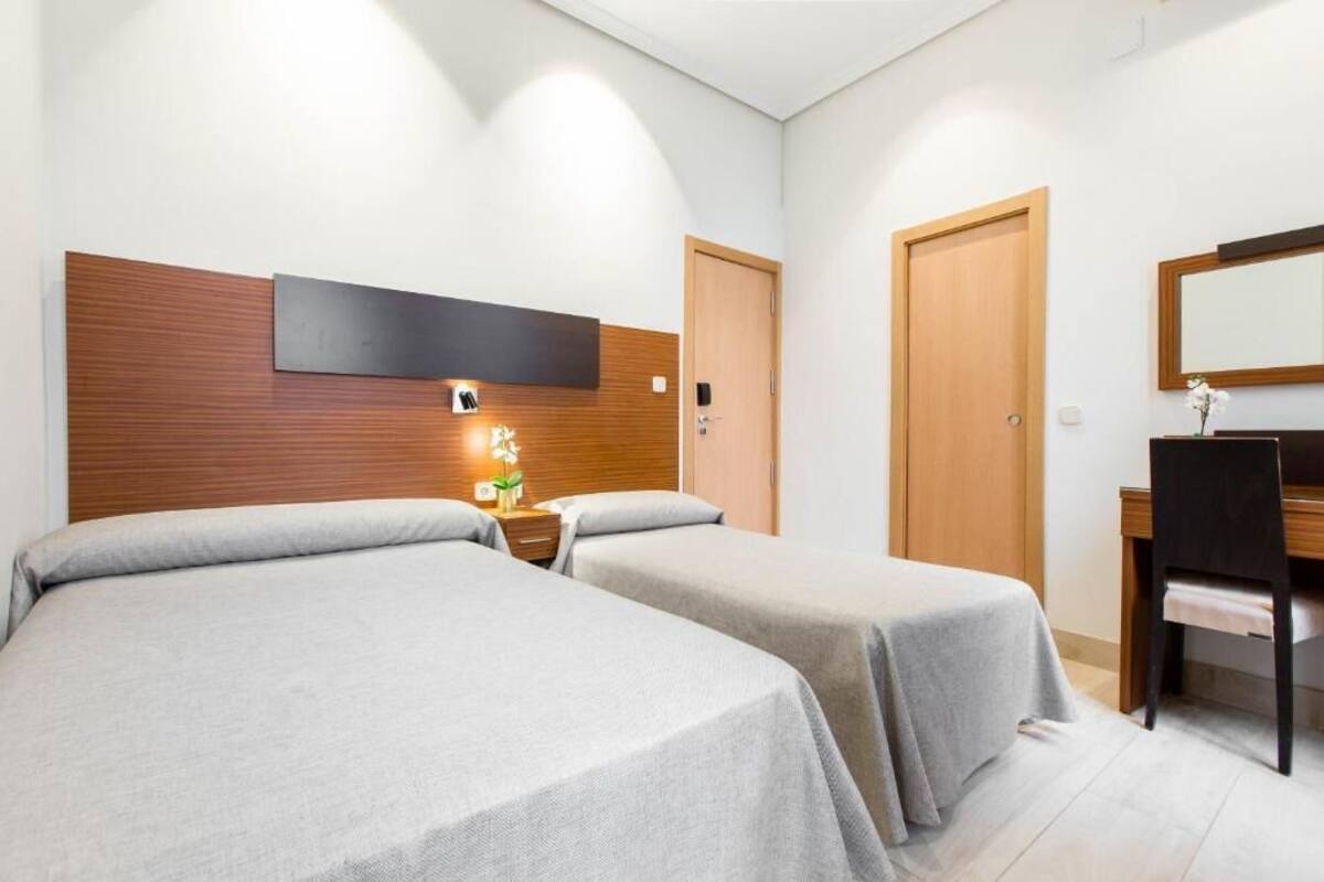 A hotel room with two beds, a wooden headboard, a bedside table with a lamp, a mirror, and a wooden door.
