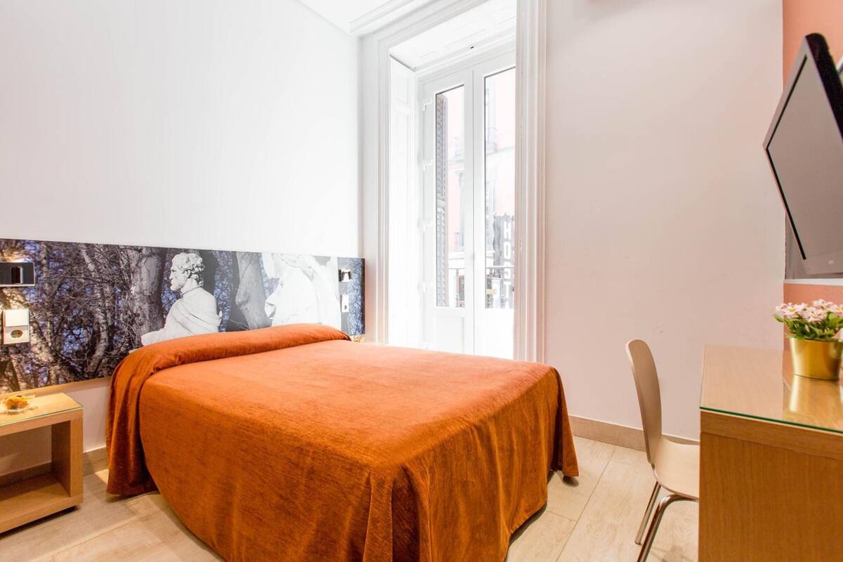A bedroom with a large bed, a desk, a chair, and a window with a view of buildings.