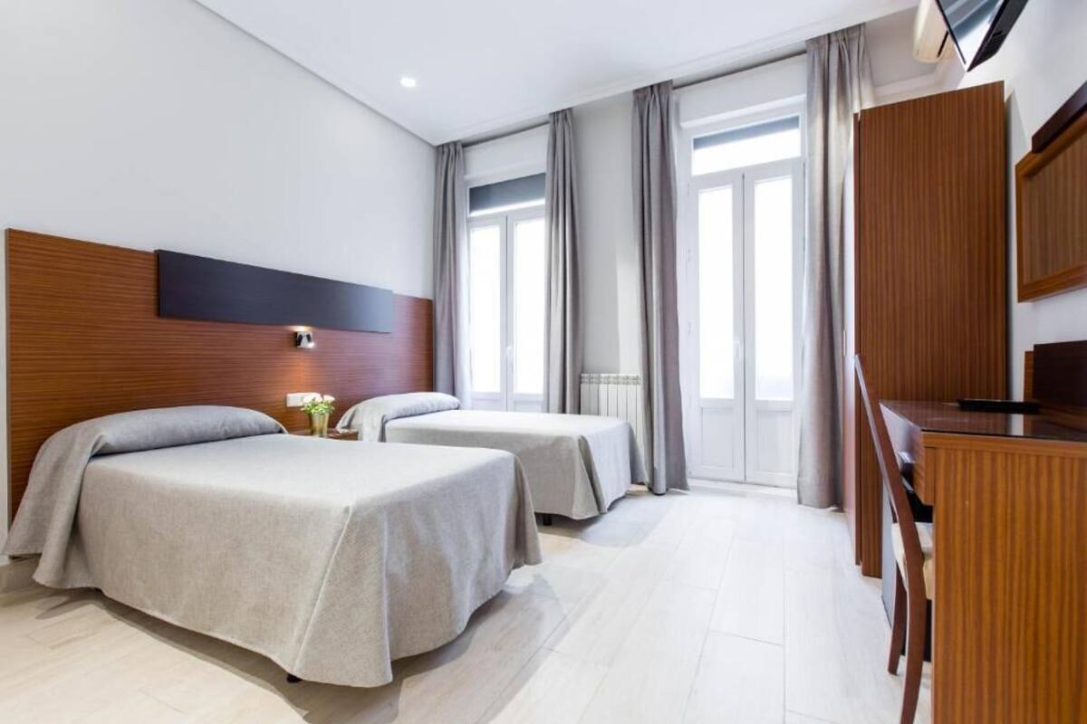 A hotel room with two beds, a wooden headboard, a desk, and a large window with curtains.