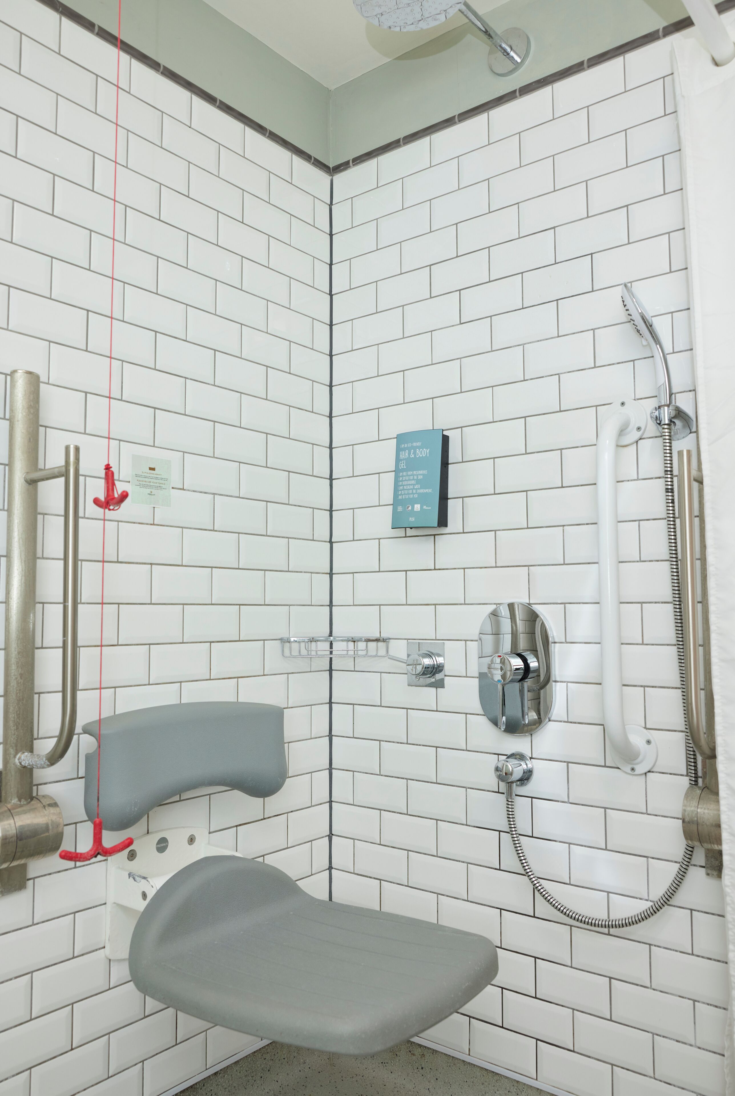A bathroom with white tiled walls, a shower with a handheld showerhead, a toilet with a safety bar, and a sign indicating the use of a specific soap.