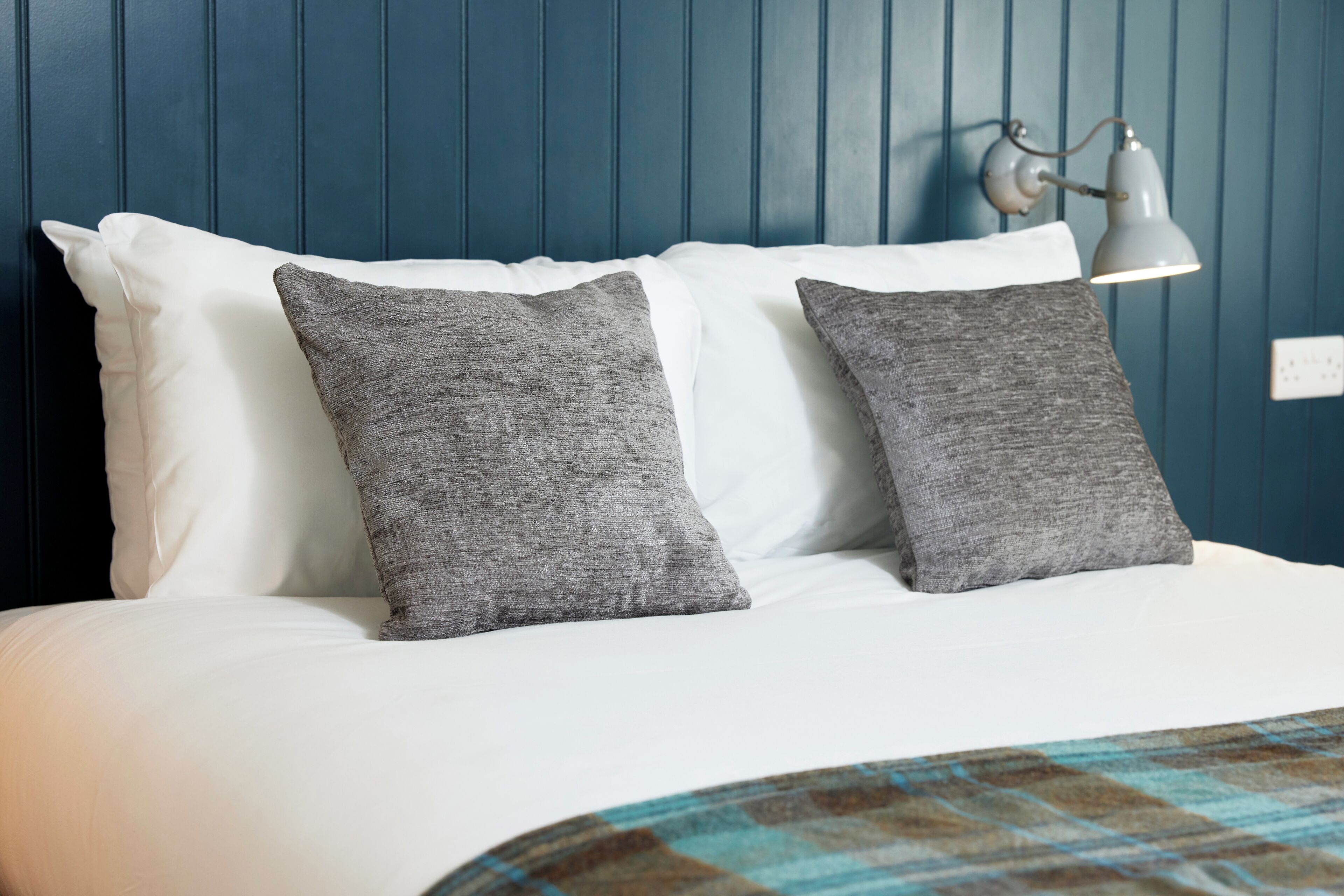 Una cama con almohadas blancas y grises, una pared azul y una lámpara fijada a la pared.