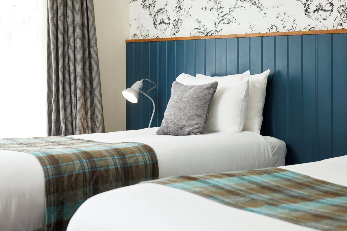 A hotel room with a bed, a bedside lamp, a window with curtains, and a floral wallpaper.