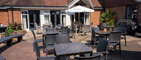 An outdoor patio area with multiple tables and chairs, a covered seating area, and a brick building with a tiled roof.