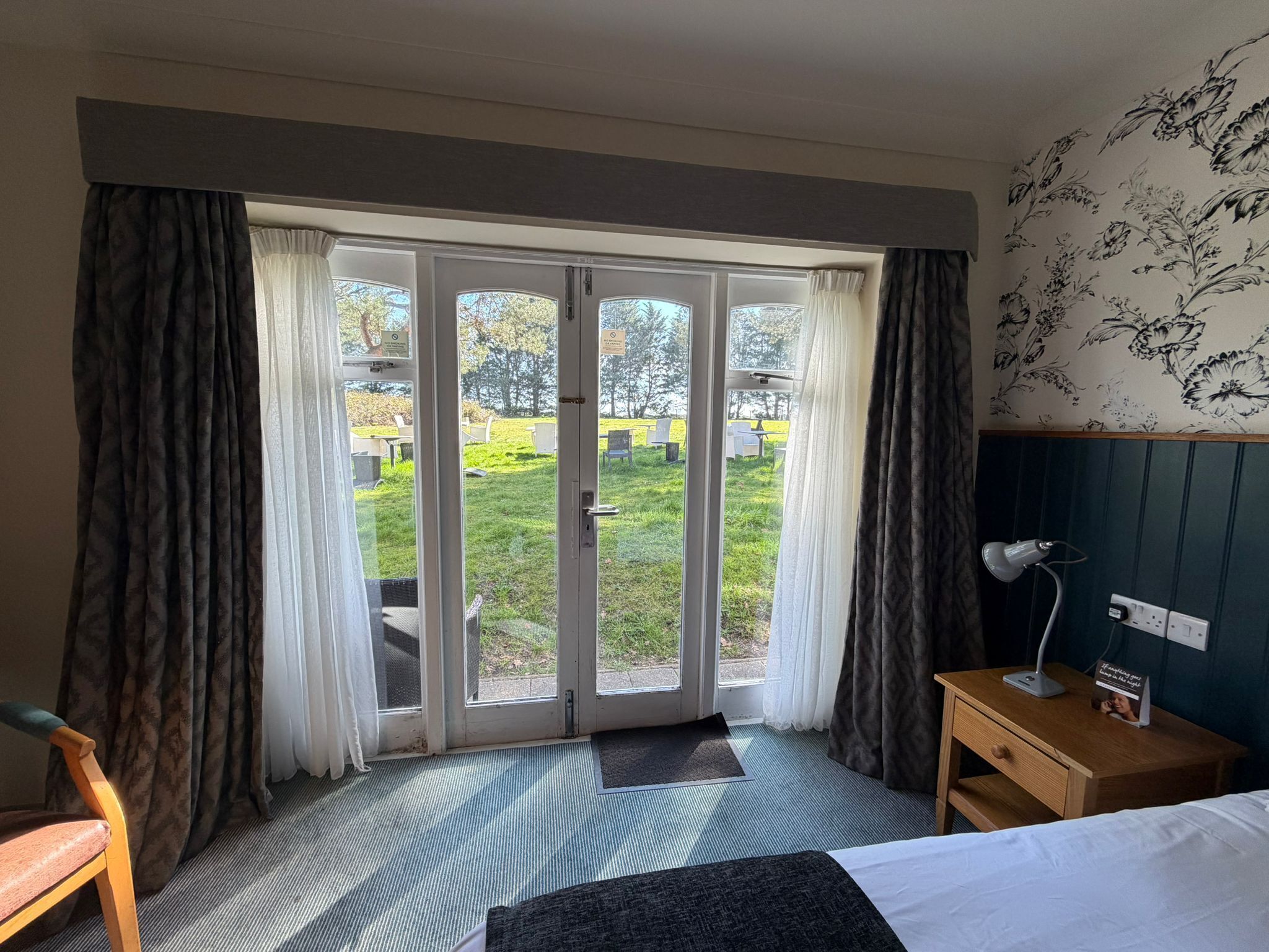 Een hotelkamer met een bed, een nachtkastje met een lamp, een stoel, en uitzicht op een groen grasveld door een glazen deur.