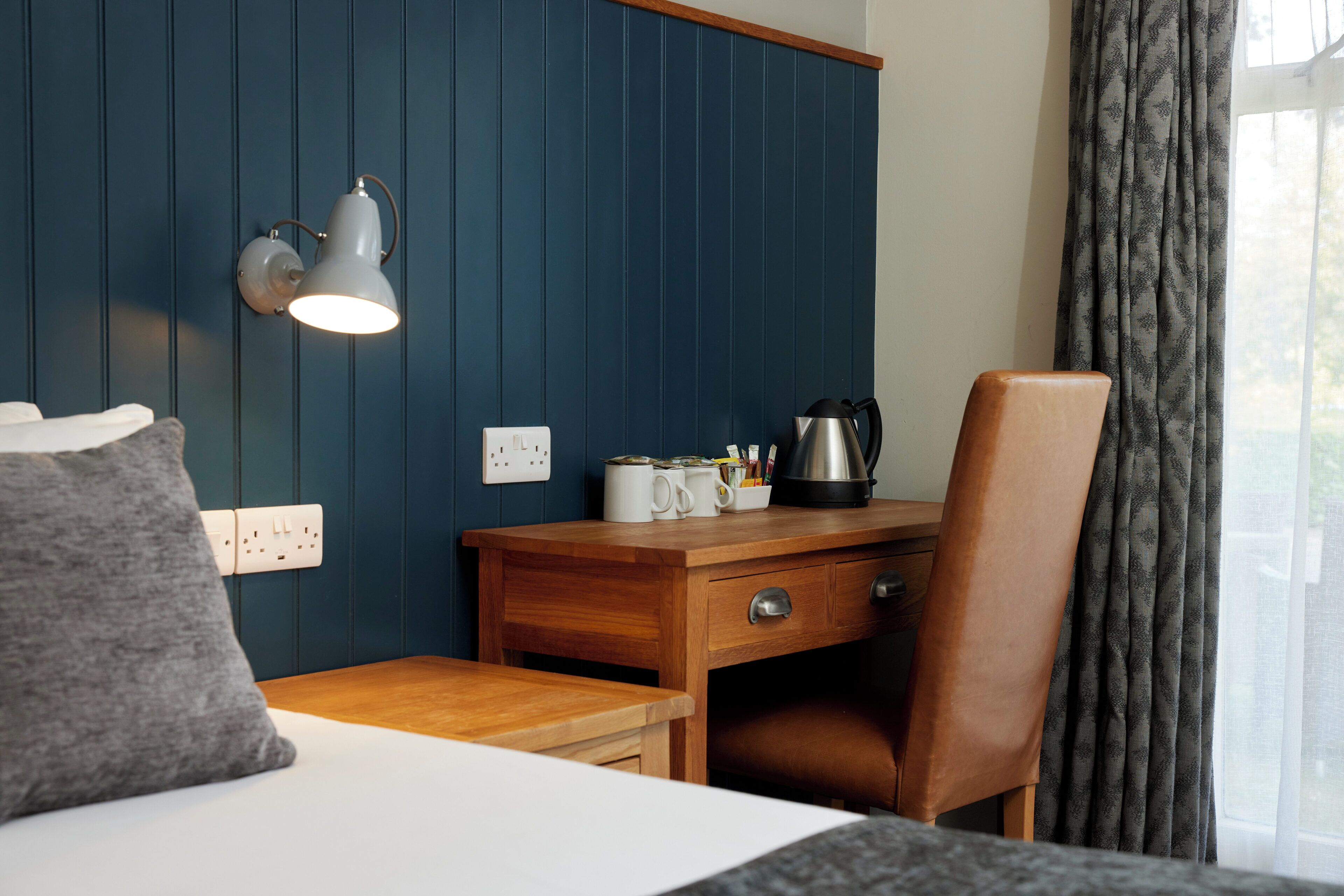 Ein Hotelzimmer mit einem Holztisch, einem Stuhl, einem Bett, einer Wandlampe und einem Fenster mit Vorhängen.