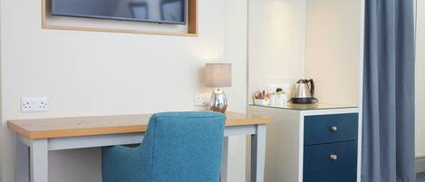 A hotel room with a wooden desk, a blue chair, a TV mounted on the wall, a bedside lamp, a nightstand with drawers, and a window with curtains.