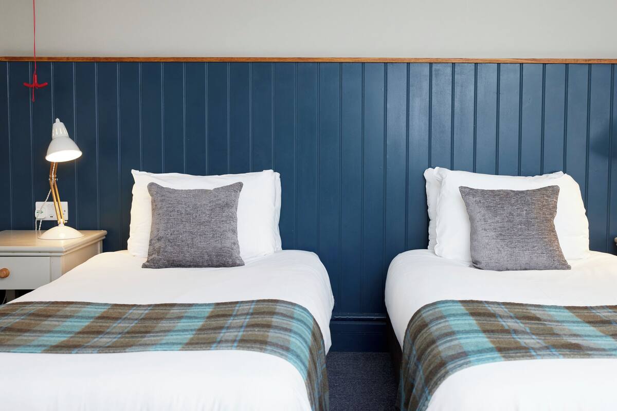 A hotel room with two beds, each with a white pillow and a grey throw pillow, flanked by nightstands with lamps.