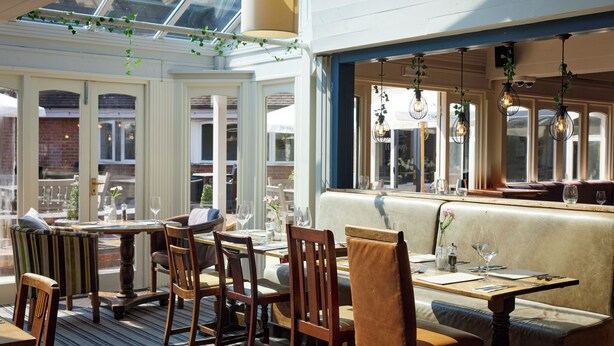 An indoor dining area with wooden tables and chairs, a bench, and hanging light fixtures. - St Leonard's Hotel by Greene King Inns (Ringwood)