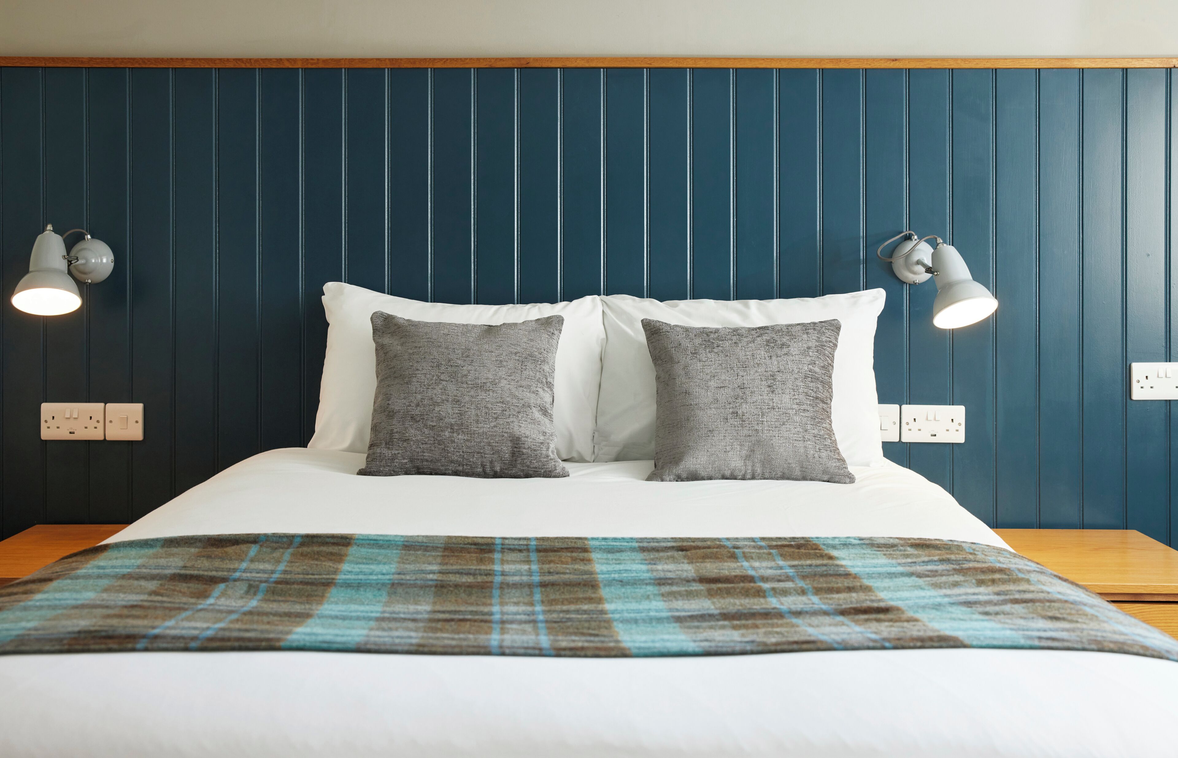 A bed with two pillows, a plaid blanket, and wall-mounted lamps.