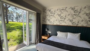 A hotel room with a bed, a nightstand, a lamp, a window with curtains, and a view of a grassy area. - St Leonard's Hotel by Greene King Inns (Ringwood)