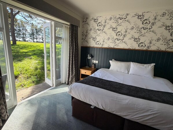 A hotel room with a bed, a nightstand, a lamp, a window with curtains, and a view of a grassy area.