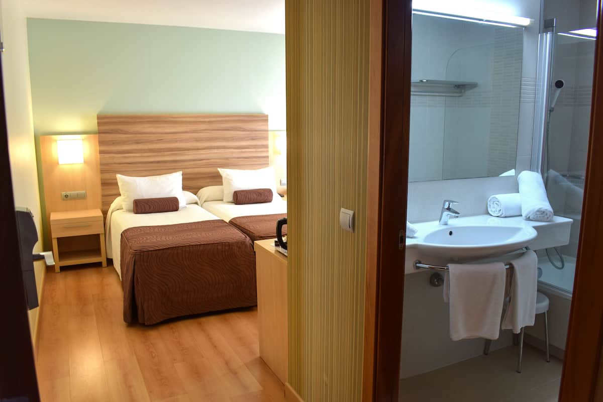 A hotel room with two beds, a wooden headboard, a bedside table with a lamp, and a bathroom with a sink, mirror, and towel rack.