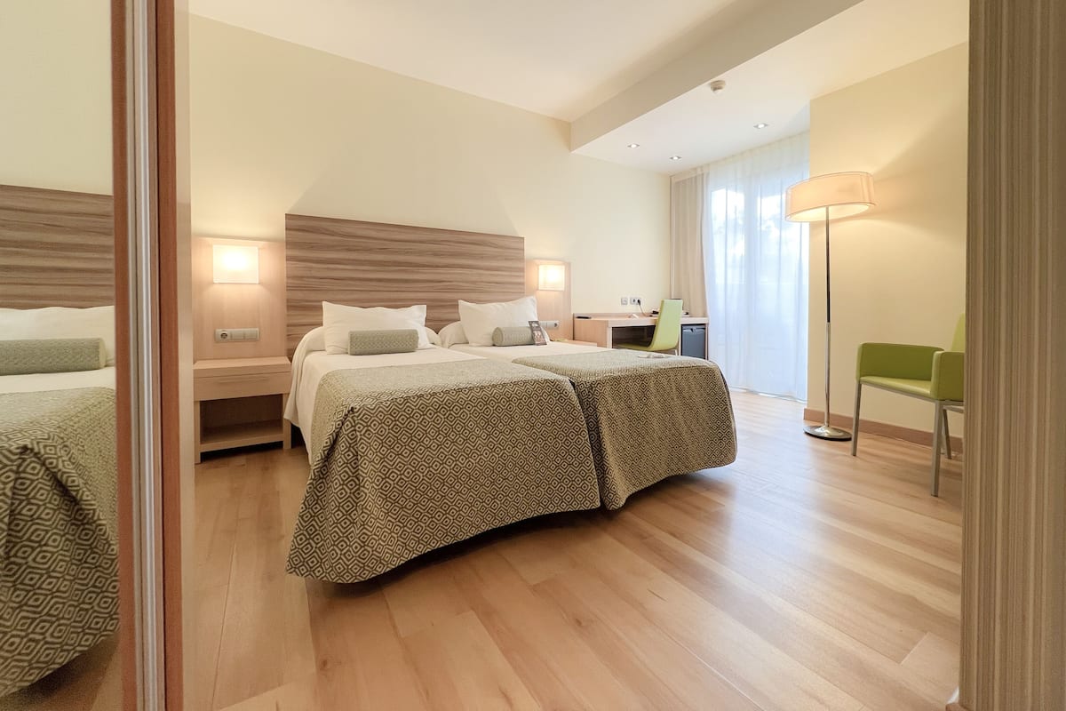 A hotel room with two beds, a desk, a chair, and a lamp.