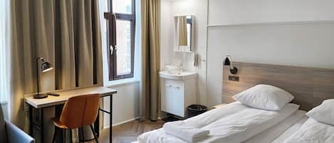 Ein Hotelzimmer mit Bett, Schreibtisch, Stuhl, einem Fenster mit Vorhängen, einem Waschbecken und einem Spiegel.