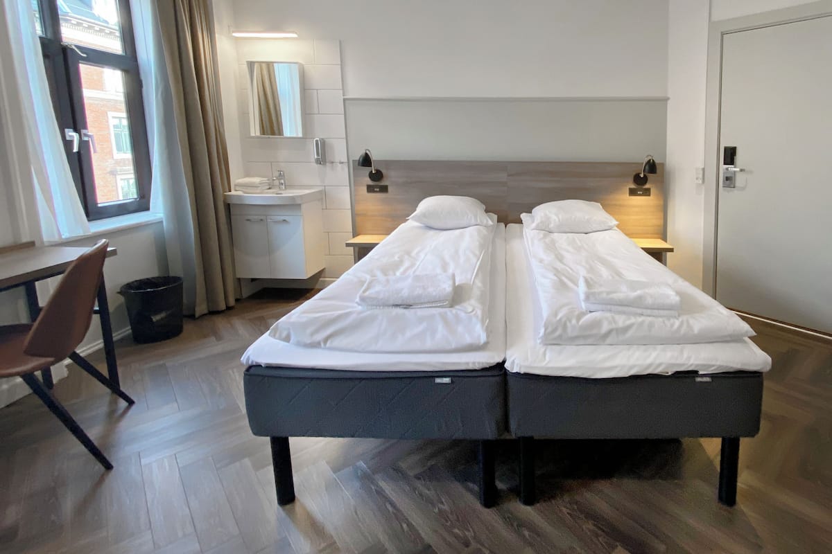Une chambre d’hôtel avec deux lits simples, un bureau, une chaise, une petite salle de bain et une fenêtre avec des rideaux.