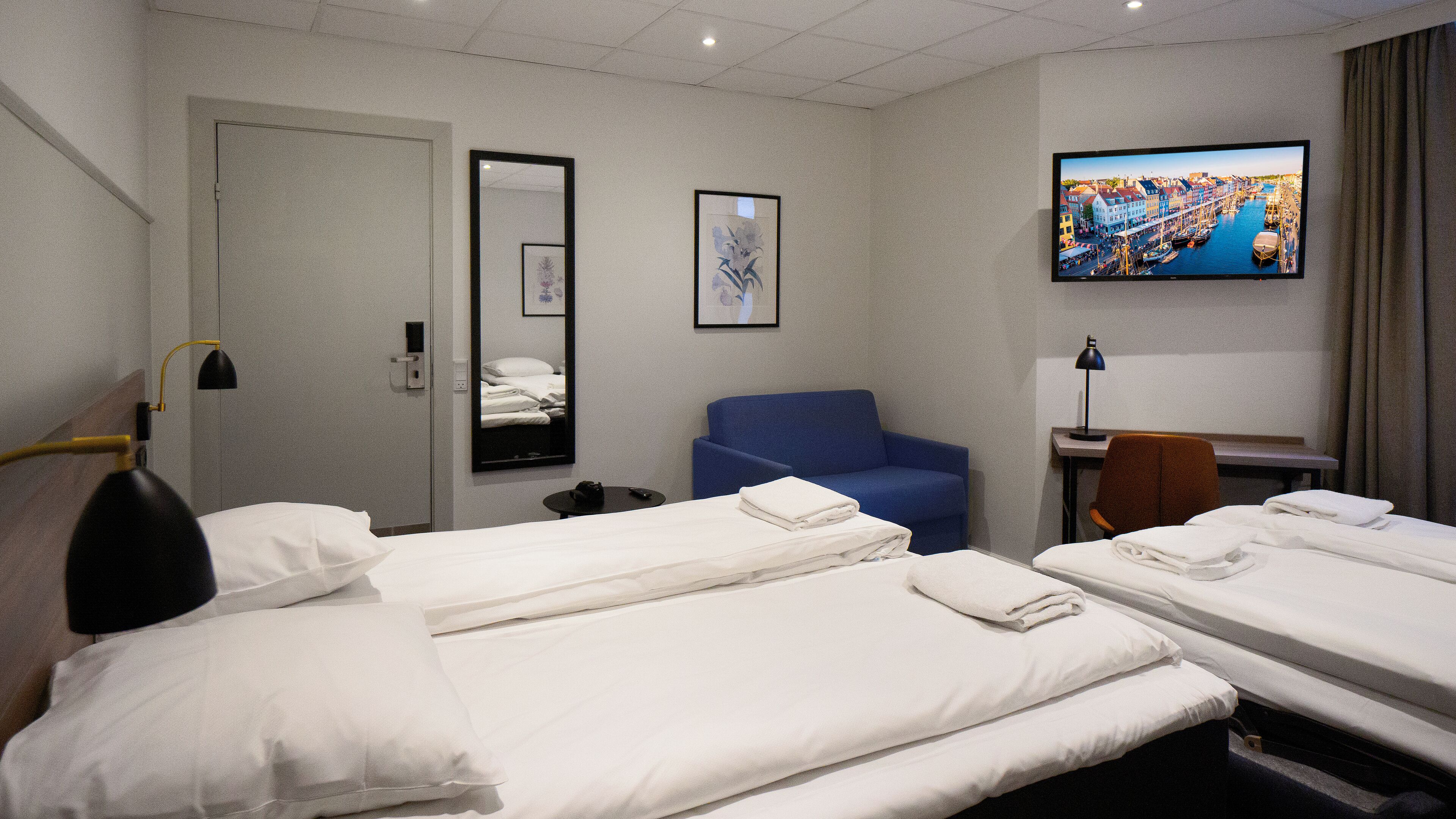 Ett hotellrum med två separata sängar, ett skrivbord med en stol, en tv, en blå soffa och en spegel.