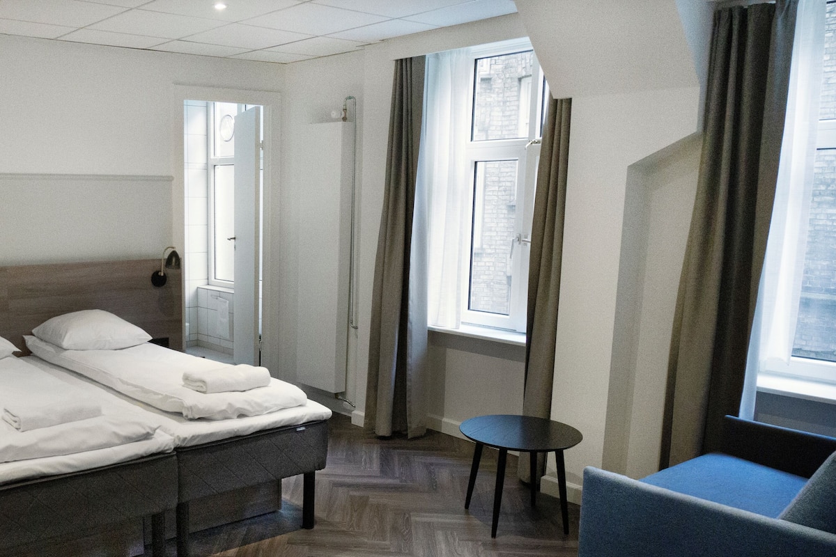 Une chambre d’hôtel avec deux lits, une petite table, une chaise et une fenêtre avec des rideaux.