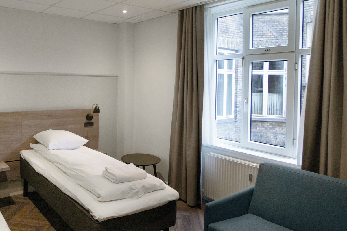 Une chambre d’hôtel avec deux lits simples, une petite table, une chaise et une fenêtre avec des rideaux.