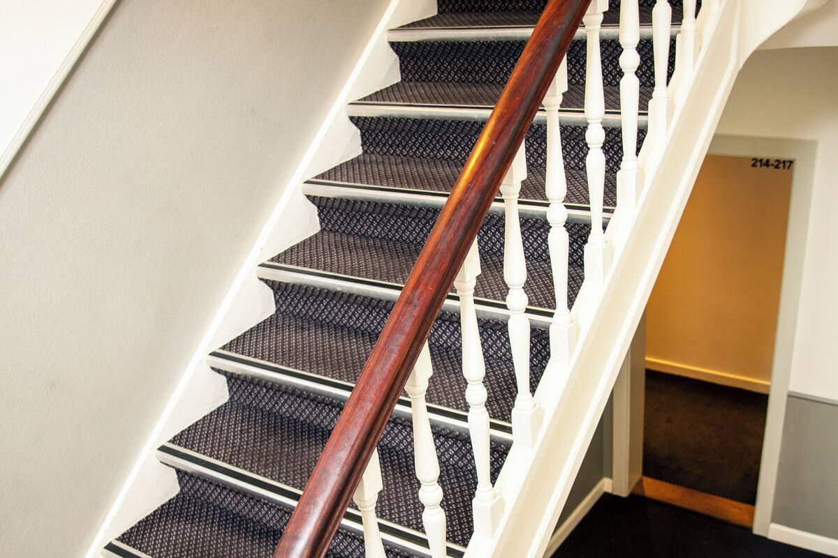 Un escalier avec une rampe en bois et des balustrades blanches, des marches recouvertes de moquette et un mur sur lequel se trouve une porte portant l’inscription « 214-217 ».